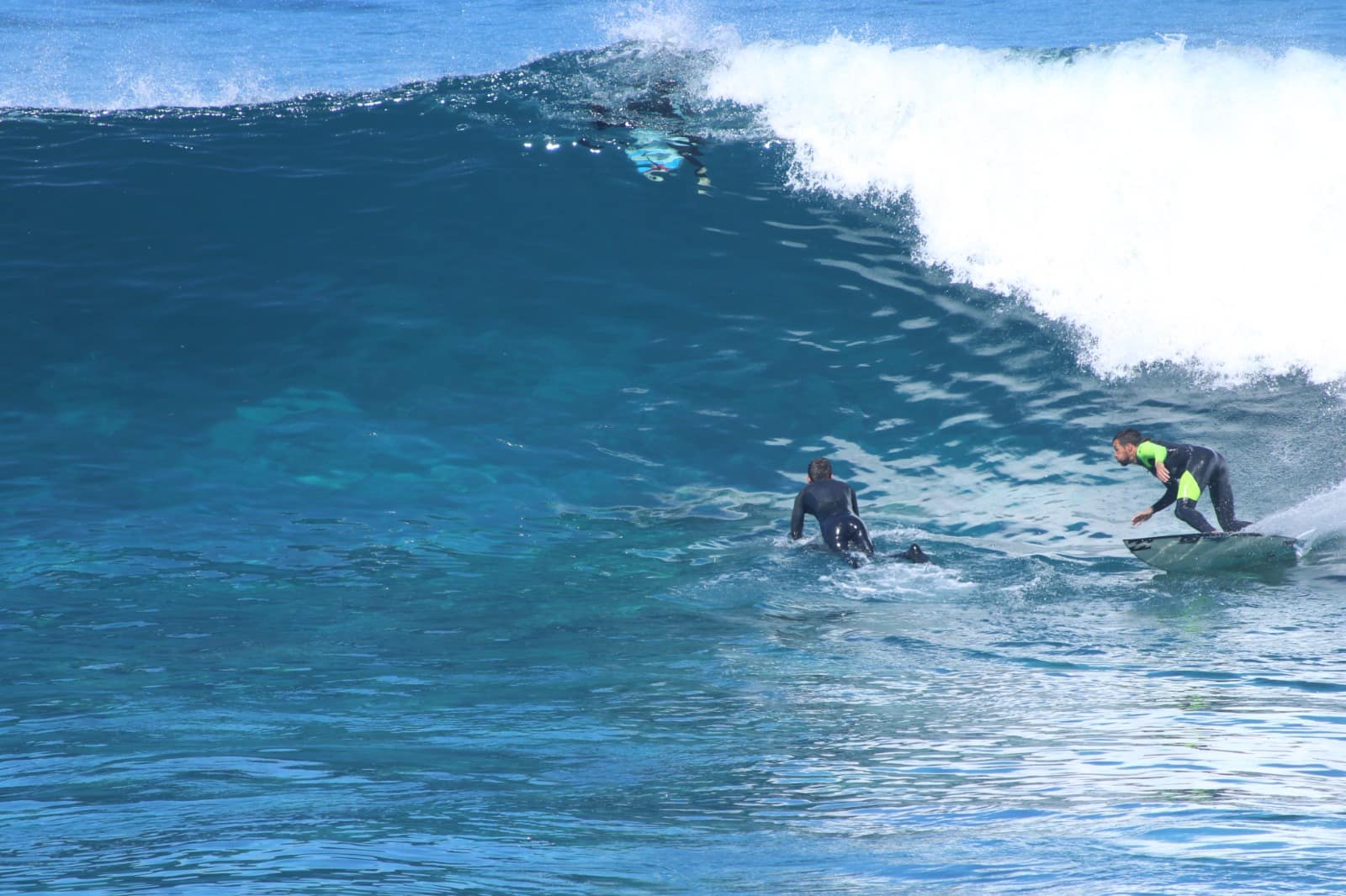 Dois surfistas na mesma onda de água azul, um deles a remar e outro de pé na prancha.